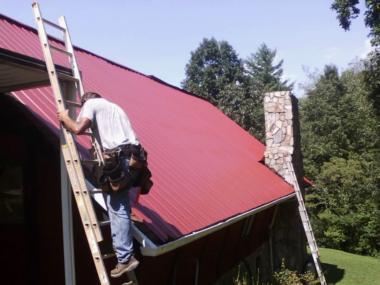 Roofer on a ladder beside a red metal roof and stone chimney — Soffit & Fascia Repair by Roofing Brothers in Franconia